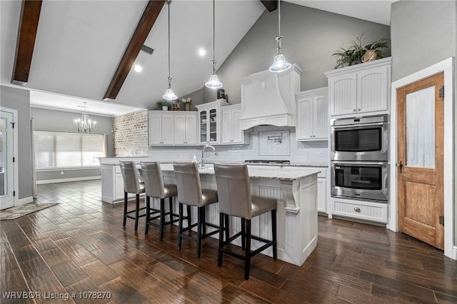 kitchen featuring decorative backsplash, white cabinets, custom range hood, beamed ceiling, and an island with sink