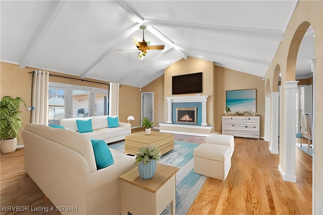 living area with light wood-style flooring, a ceiling fan, lofted ceiling with beams, a warm lit fireplace, and arched walkways