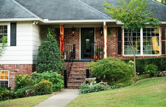 view of exterior entry featuring a yard, roof with shingles, and brick siding