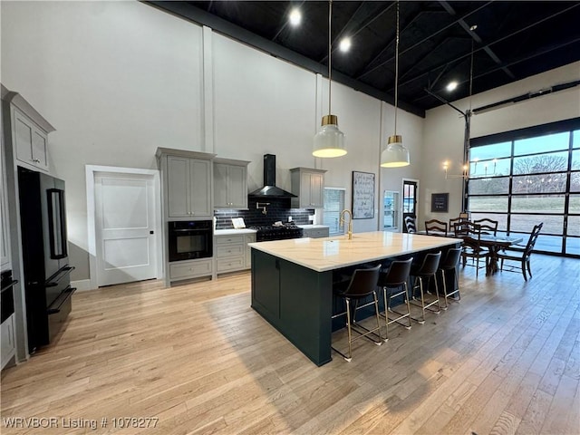 kitchen with a towering ceiling, black appliances, wall chimney range hood, a center island with sink, and gray cabinets