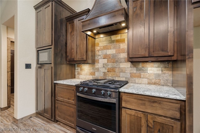 kitchen with stainless steel gas range oven, light stone counters, custom range hood, light wood-style floors, and tasteful backsplash