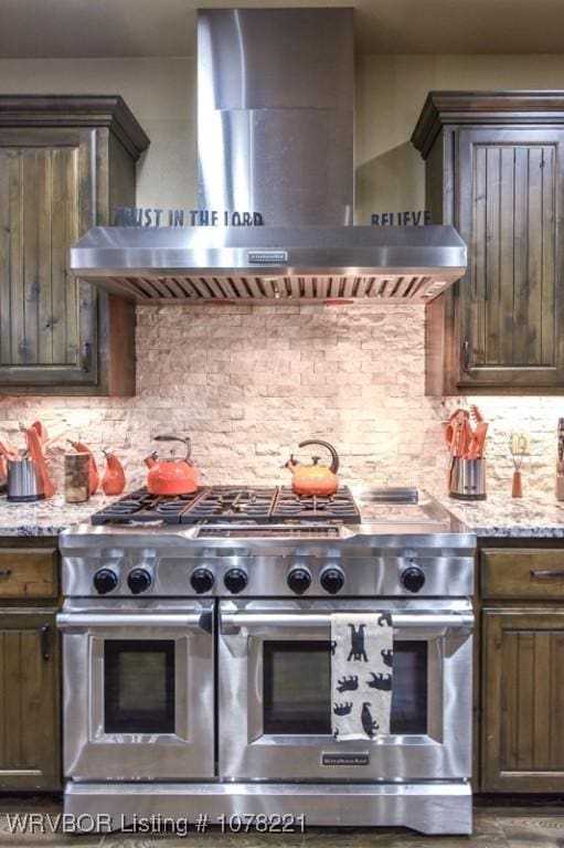 kitchen with decorative backsplash, range with two ovens, light stone countertops, and wall chimney range hood