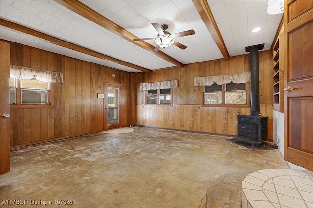 unfurnished living room with beam ceiling, a ceiling fan, a wood stove, wooden walls, and concrete floors