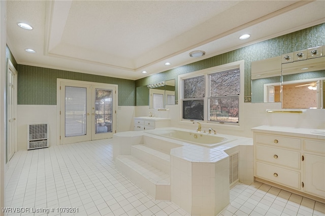 bathroom with a tray ceiling, french doors, a garden tub, two vanities, and wallpapered walls