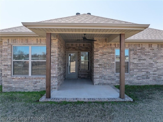 doorway to property with brick siding, a shingled roof, ceiling fan, a yard, and a patio area