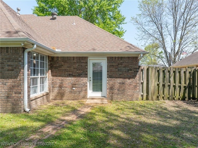 doorway to property featuring brick siding, fence, a yard, and a shingled roof