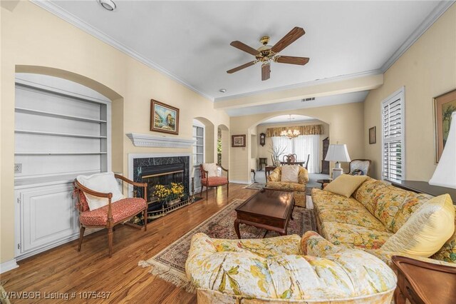 living room with built in shelves, ceiling fan with notable chandelier, crown molding, a premium fireplace, and hardwood / wood-style floors