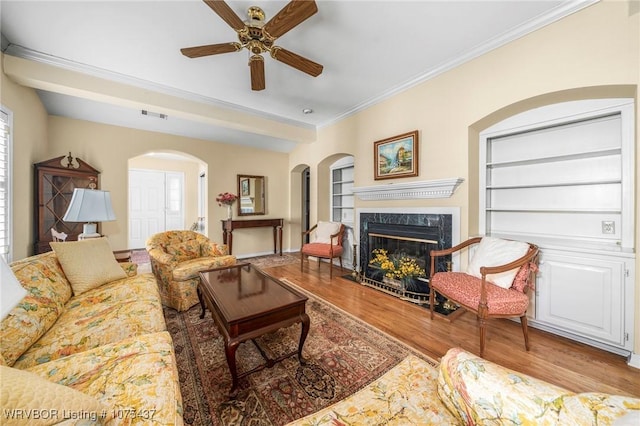 living room with ceiling fan, a premium fireplace, built in features, hardwood / wood-style flooring, and ornamental molding