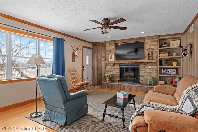 living room with a textured ceiling, ceiling fan, a stone fireplace, light wood-style floors, and crown molding