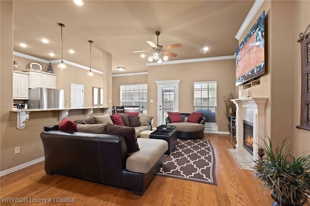 living room featuring a fireplace with flush hearth, light wood-type flooring, baseboards, a ceiling fan, and ornamental molding