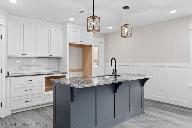 kitchen with dark stone counters, an island with sink, hanging light fixtures, white cabinetry, and a sink