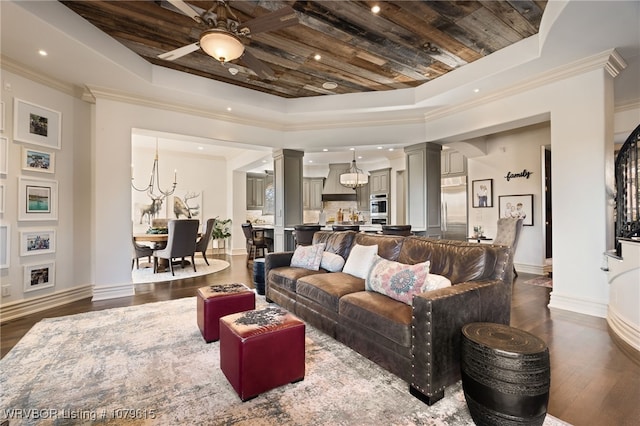 living room with ornamental molding, ceiling fan with notable chandelier, a tray ceiling, dark wood-style floors, and ornate columns