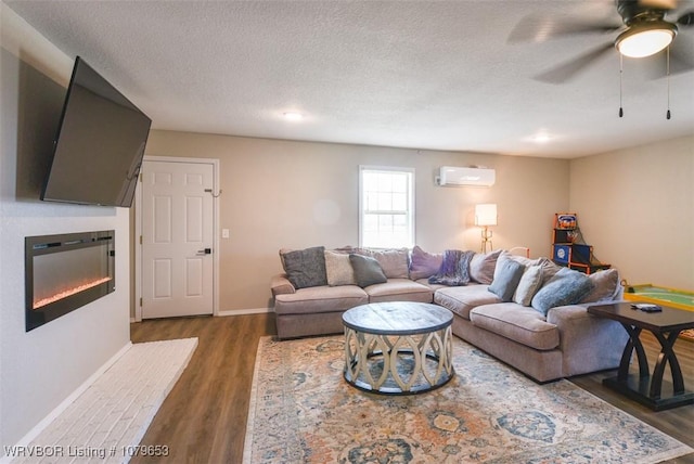 living area with a textured ceiling, wood finished floors, a glass covered fireplace, a wall unit AC, and ceiling fan