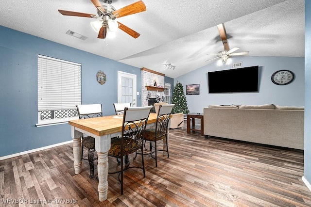 dining space with vaulted ceiling with beams, a textured ceiling, a large fireplace, ceiling fan, and hardwood / wood-style floors