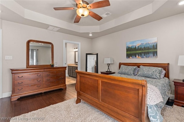 bedroom with visible vents, connected bathroom, dark wood-style floors, a raised ceiling, and a ceiling fan