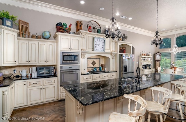 kitchen featuring an island with sink, stainless steel appliances, dark stone counters, hanging light fixtures, and sink