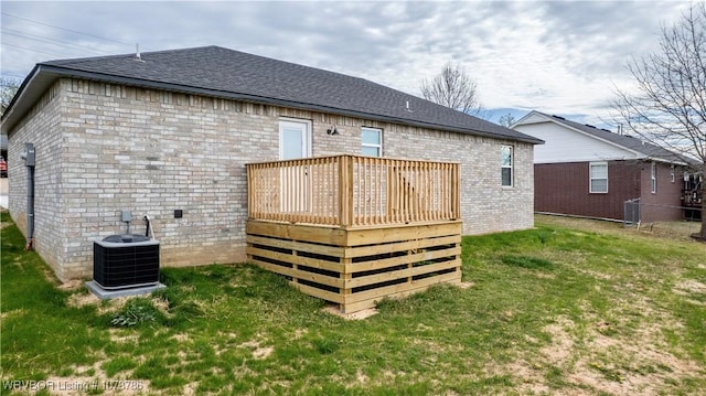 back of property featuring central air condition unit, a wooden deck, brick siding, a lawn, and a shingled roof