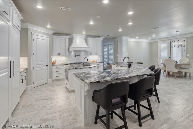 kitchen with a large island with sink, sink, custom range hood, light stone counters, and white cabinetry