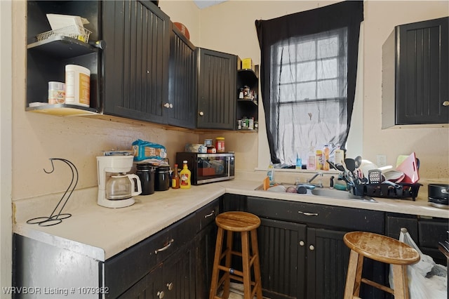 kitchen featuring open shelves, stainless steel microwave, a sink, light countertops, and dark cabinets