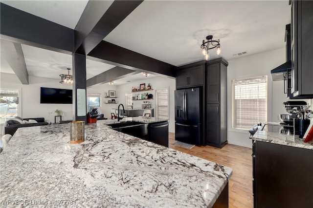 kitchen with black refrigerator with ice dispenser, beamed ceiling, light stone counters, and stainless steel dishwasher