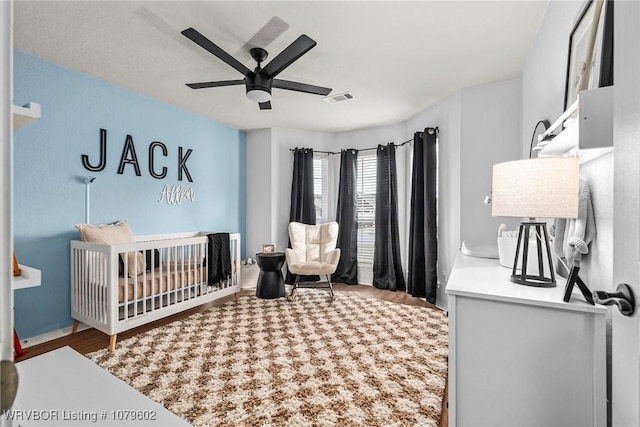 bedroom featuring ceiling fan, visible vents, a nursery area, and wood finished floors