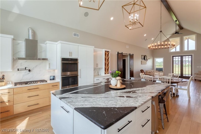 kitchen with appliances with stainless steel finishes, white cabinetry, beam ceiling, and wall chimney range hood