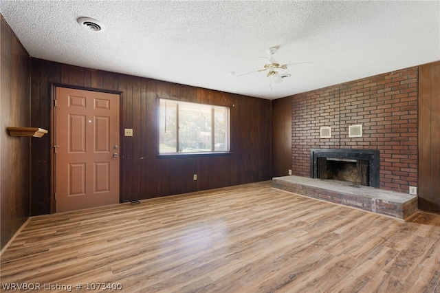 unfurnished living room with ceiling fan, a brick fireplace, a textured ceiling, wooden walls, and light wood-type flooring