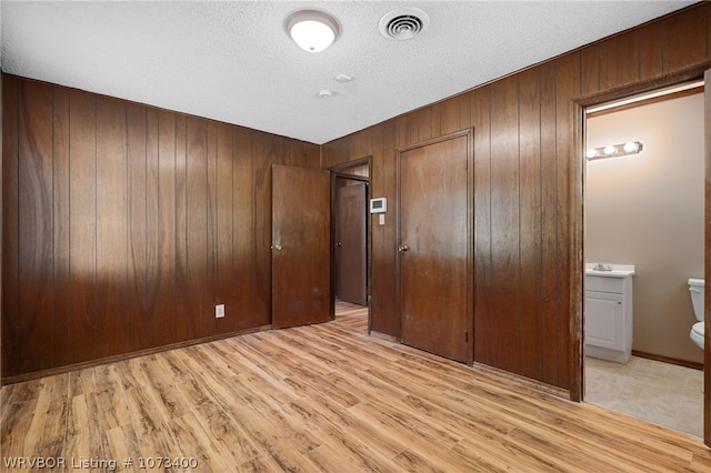 unfurnished bedroom featuring light wood-type flooring, a textured ceiling, connected bathroom, a closet, and wood walls