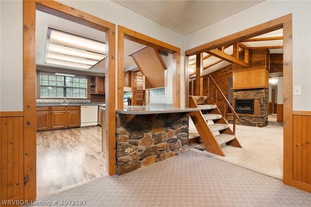 kitchen featuring dishwasher, sink, a stone fireplace, wood walls, and light colored carpet