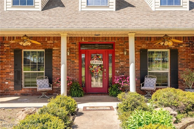 doorway to property featuring brick siding, a porch, a ceiling fan, and roof with shingles