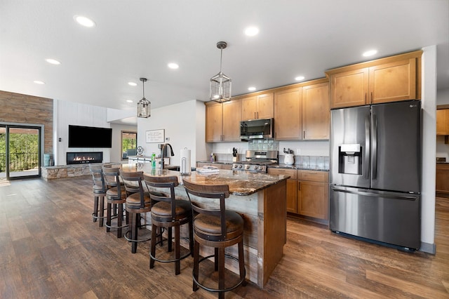 kitchen featuring dark hardwood / wood-style floors, appliances with stainless steel finishes, a center island with sink, a stone fireplace, and dark stone counters