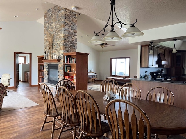 dining space featuring ceiling fan, a stone fireplace, a textured ceiling, hardwood / wood-style floors, and vaulted ceiling