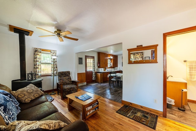 living area featuring a baseboard heating unit, a wood stove, wood-type flooring, and ceiling fan