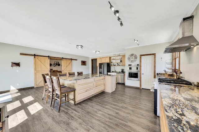 kitchen featuring a barn door, stainless steel appliances, a kitchen breakfast bar, wall chimney range hood, and light brown cabinetry