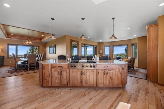 kitchen featuring stainless steel gas stovetop, hanging light fixtures, light stone counters, a tray ceiling, and a kitchen island with sink