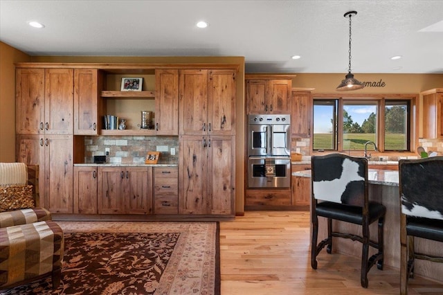 kitchen with stainless steel double oven, decorative backsplash, hanging light fixtures, a breakfast bar area, and light hardwood / wood-style floors