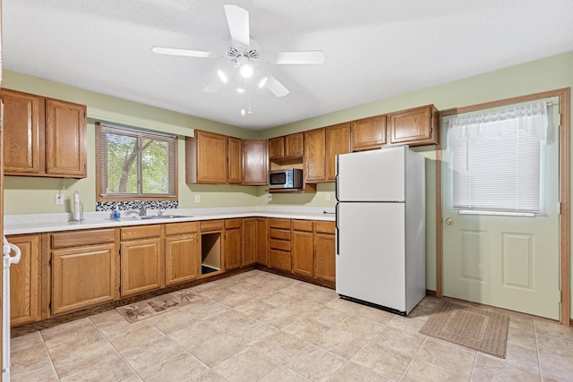 kitchen featuring brown cabinets, a sink, stainless steel microwave, freestanding refrigerator, and light countertops