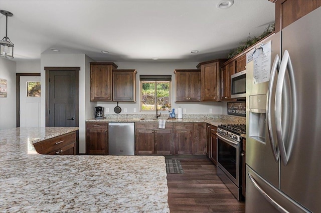 kitchen with appliances with stainless steel finishes, dark wood-type flooring, hanging light fixtures, light stone counters, and dark brown cabinetry