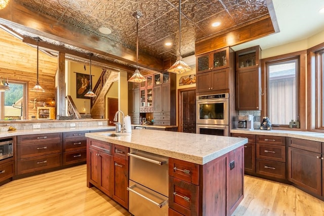 kitchen with appliances with stainless steel finishes, an island with sink, pendant lighting, light wood-type flooring, and sink