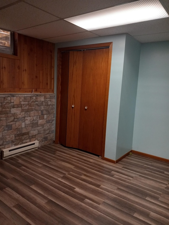 basement with dark wood-type flooring, wood walls, a drop ceiling, and baseboard heating