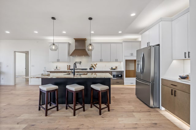 kitchen featuring light stone countertops, stainless steel fridge, custom exhaust hood, white cabinetry, and an island with sink