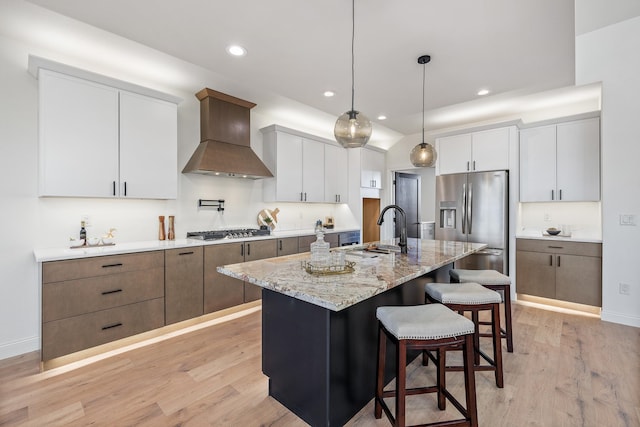 kitchen featuring pendant lighting, a center island with sink, wall chimney range hood, white cabinetry, and stainless steel appliances