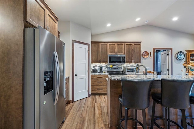 kitchen featuring light stone countertops, stainless steel appliances, vaulted ceiling, decorative backsplash, and light wood-type flooring