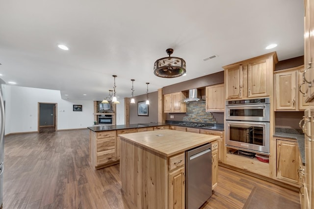 kitchen with light brown cabinetry, appliances with stainless steel finishes, wall chimney range hood, decorative light fixtures, and a kitchen island