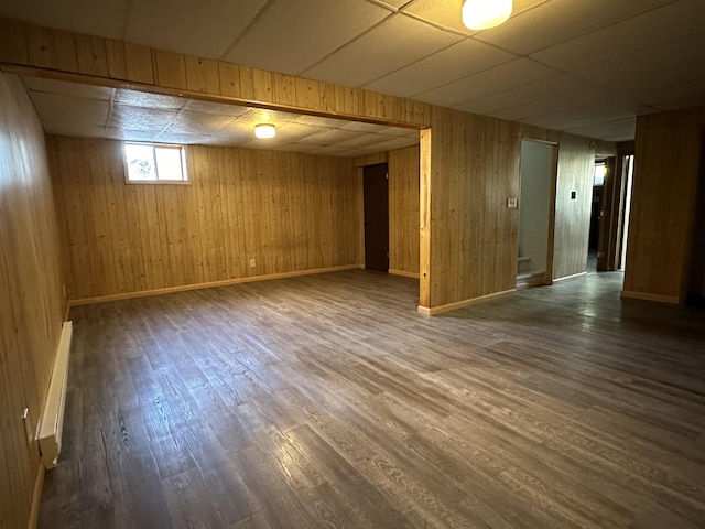 basement with a paneled ceiling, dark hardwood / wood-style floors, wood walls, and a baseboard radiator