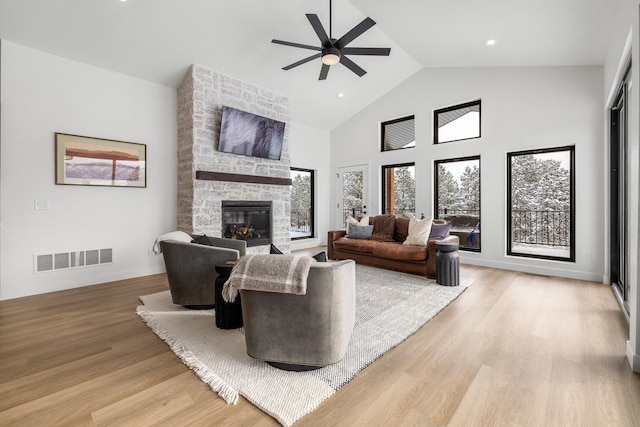 living room with ceiling fan, a stone fireplace, high vaulted ceiling, and light wood-type flooring