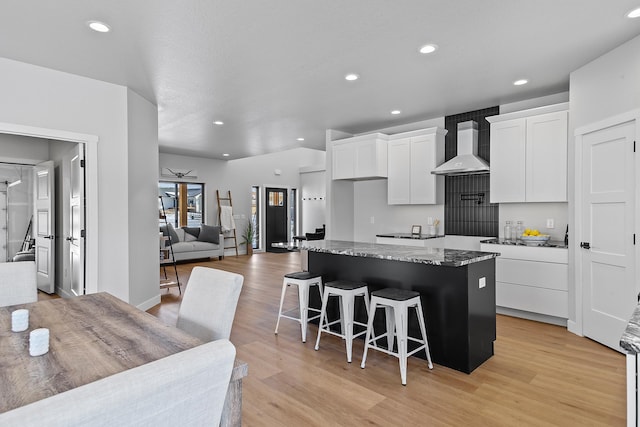 kitchen with wall chimney exhaust hood, a kitchen bar, white cabinetry, a kitchen island, and dark stone counters