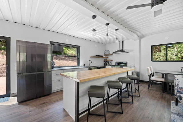 kitchen featuring wall chimney range hood, appliances with stainless steel finishes, white cabinets, a kitchen bar, and wood counters