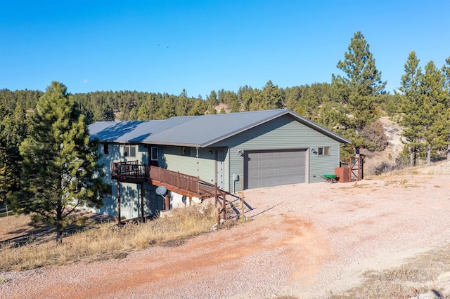 view of front facade with an attached garage, dirt driveway, a view of trees, and a wooden deck