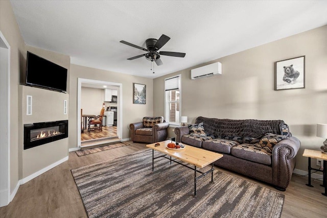 living area with a glass covered fireplace, ceiling fan, a wall unit AC, and wood finished floors